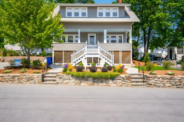 Gray and white two-story house with front porch, white railings, stone foundation, and landscaped yard