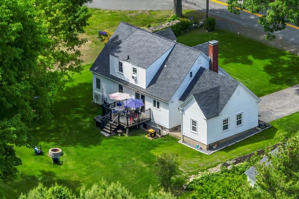 Aerial view of white farmhouse with gray roof, deck with umbrellas, large green lawn, and surrounding trees