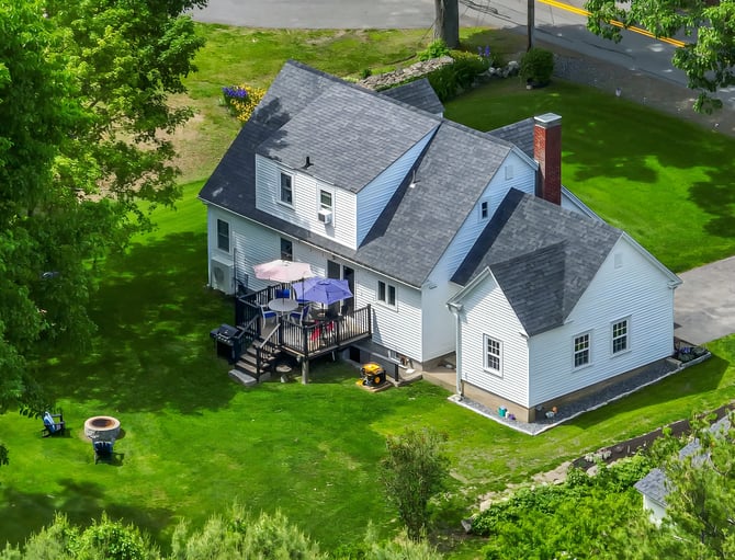 Aerial view of white two-story house with gray roof, deck with umbrellas, large lawn, and surrounding trees